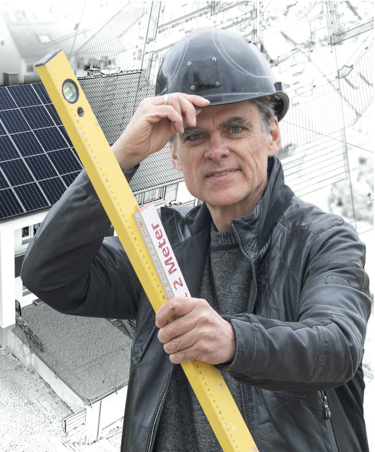 Ronald Meyer mit schwarzem Bauhelm und Wasserwaage vor einem Haus mit Solardach und einer Planskizze